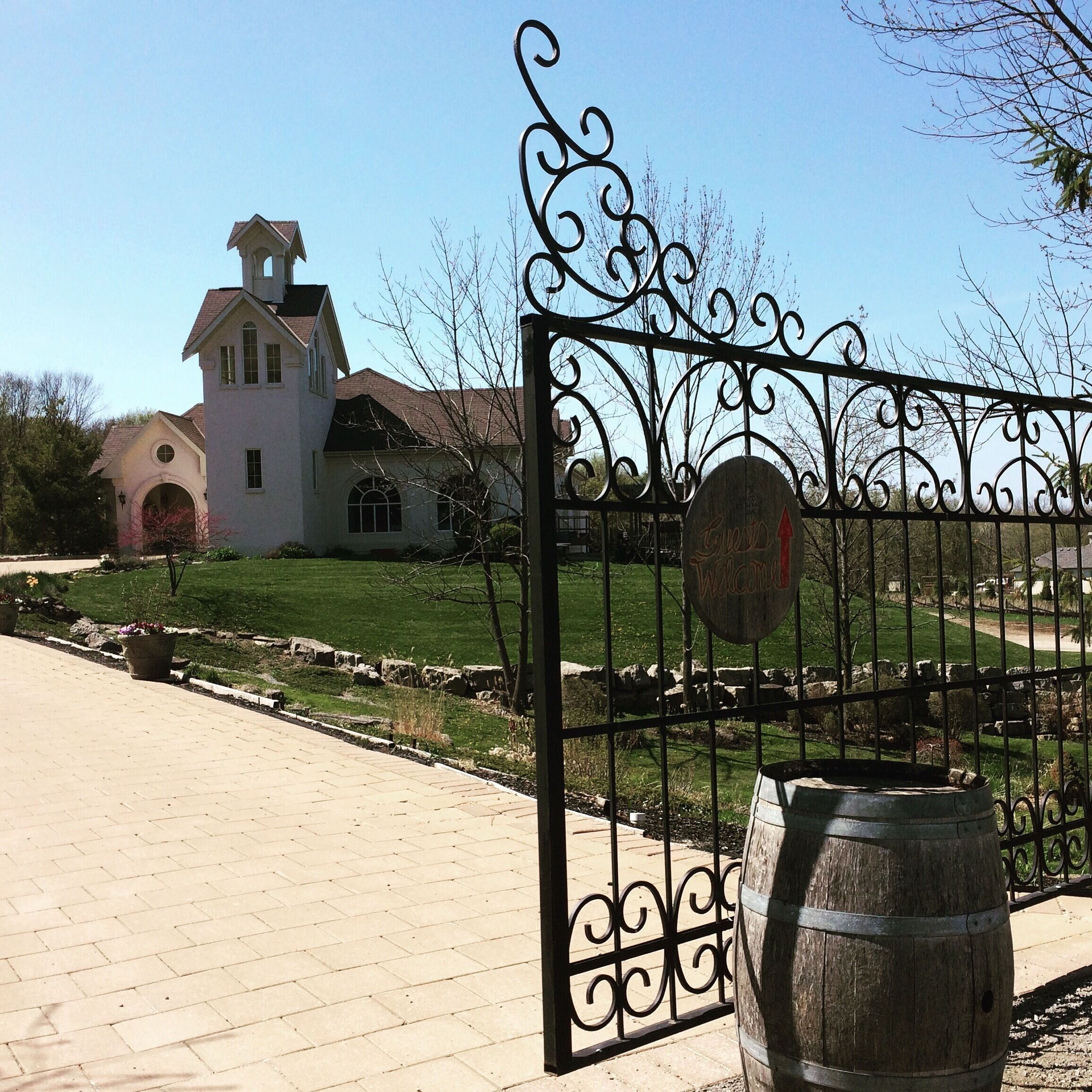 A gated entrance with a metal gate and a wooden barrel, leading to a white church with a bell tower.