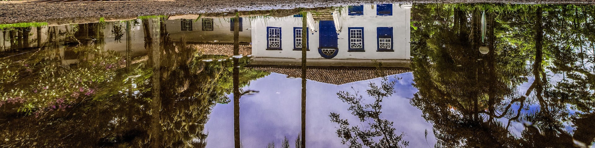 A white building with blue doors and windows, surrounded by lush greenery and a reflective pond.