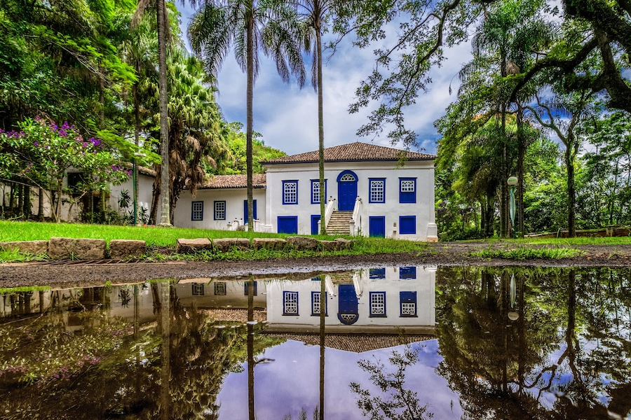 Un edificio blanco con puertas y ventanas azules, rodeado de abundante vegetación y un estanque que refleja el paisaje.