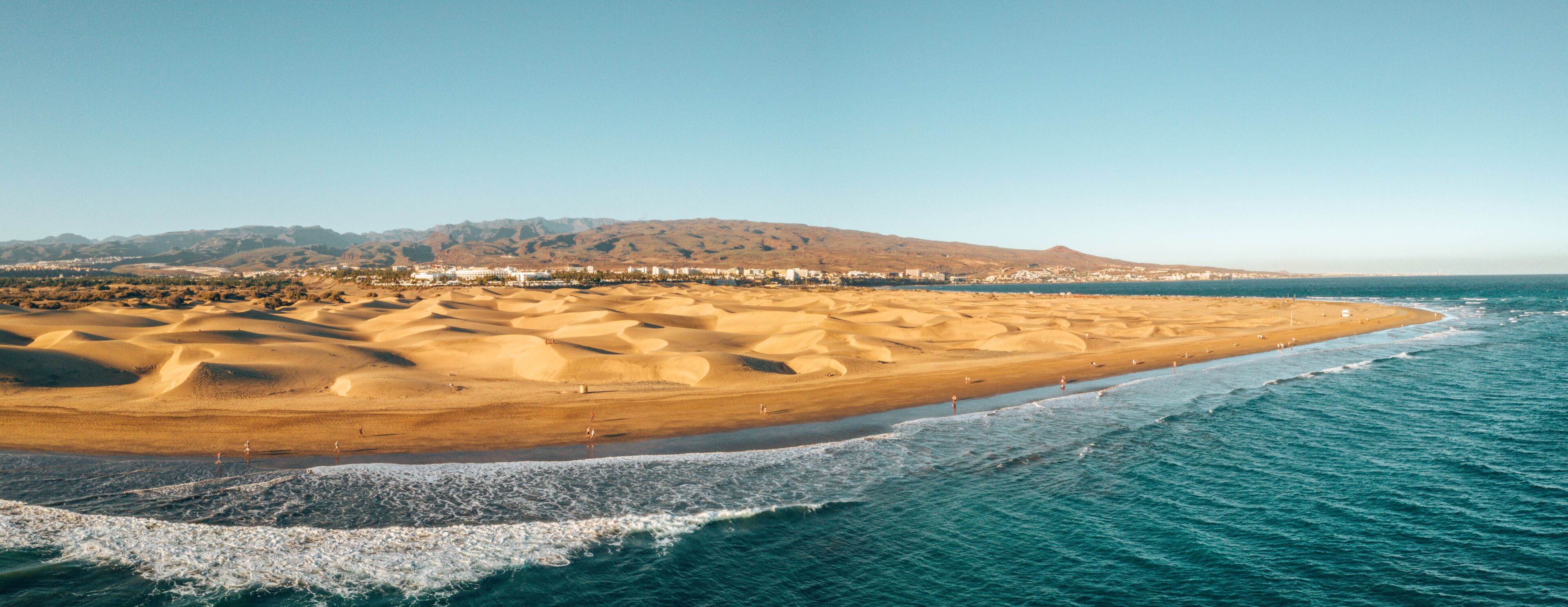 A coastal landscape with a sandy beach, ocean waves, and a distant mountain range.