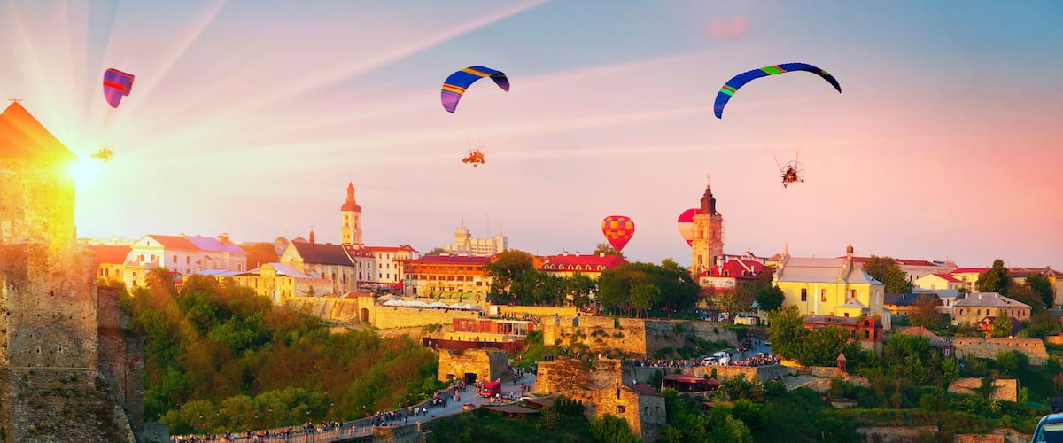 A historic cityscape with a prominent castle, colorful hot air balloons, and a clear sky.