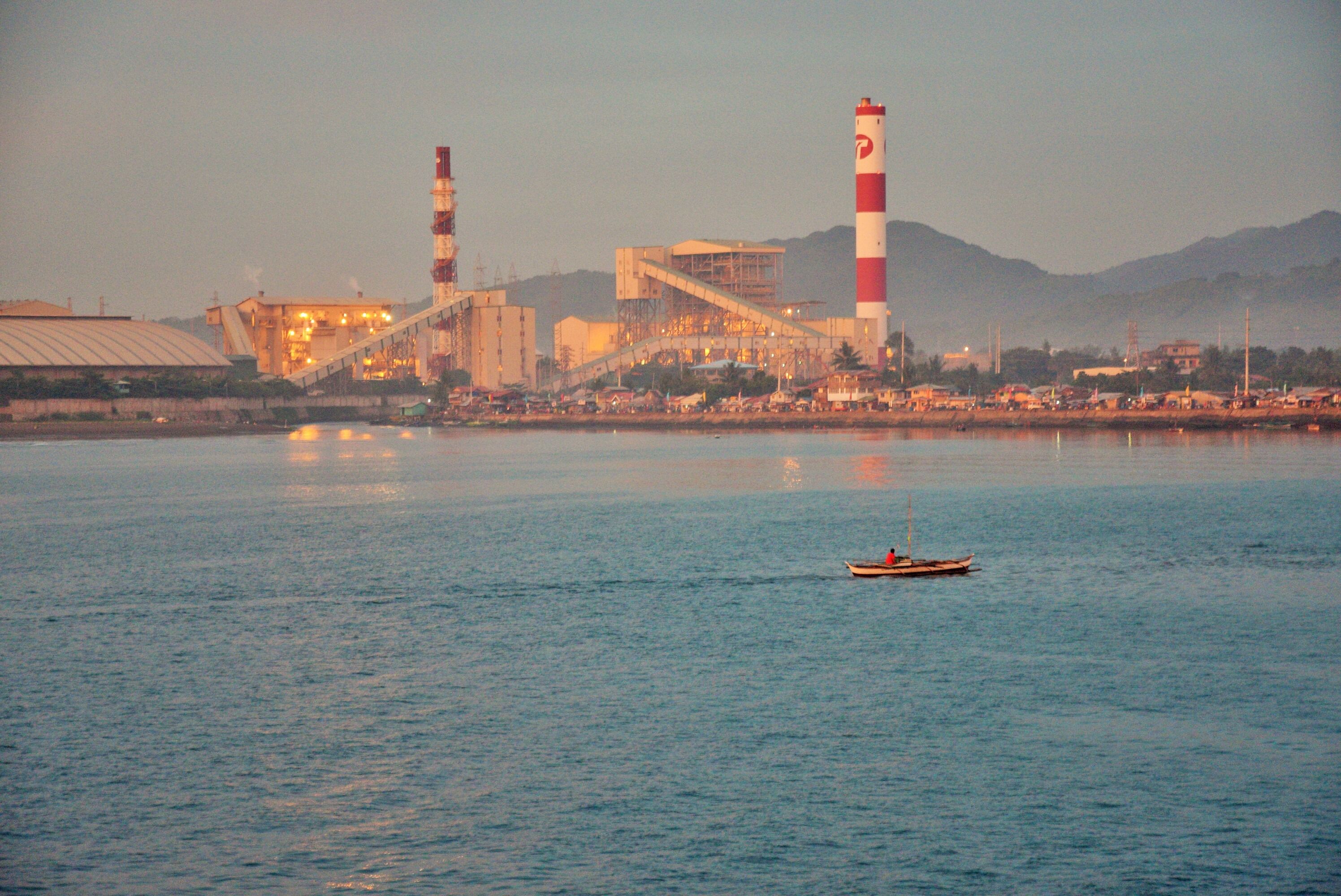 A coastal industrial facility with two tall chimneys, a large building, and a calm body of water with a small boat.
