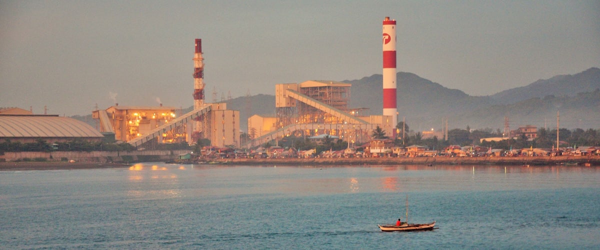A coastal industrial facility with two tall chimneys, a large building, and a calm body of water with a small boat.