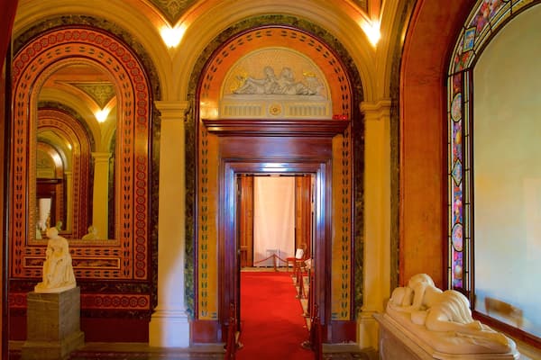 An ornate hallway with red carpet, classical statues, and stained glass windows.