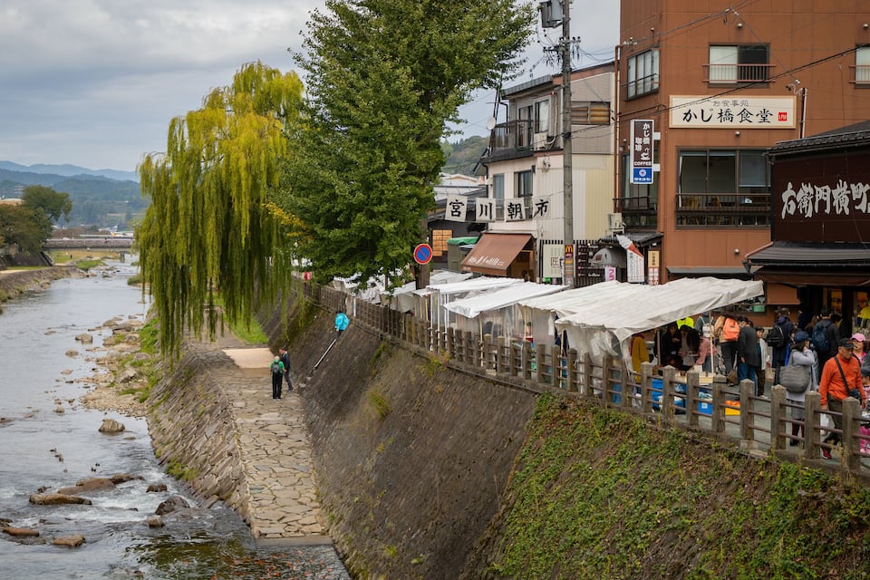 Un paseo a orillas del río con un muro de piedra, sauces y una multitud de gente cerca de tiendas y restaurantes.