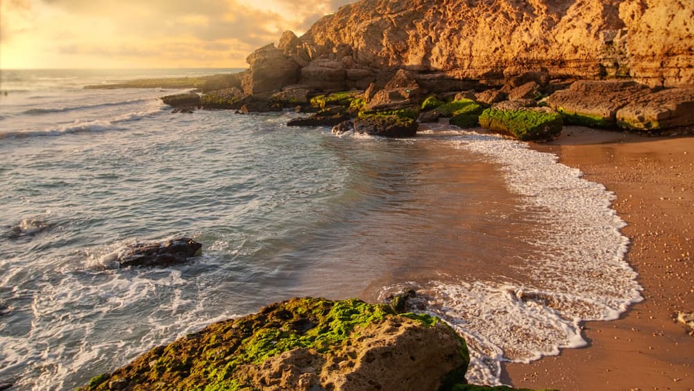 A coastal landscape with rocky cliffs, green moss-covered rocks, and waves crashing onto the sandy beach.
