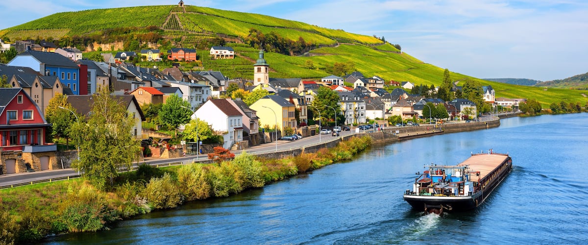 A river town with a boat, houses, and a hill with vineyards.