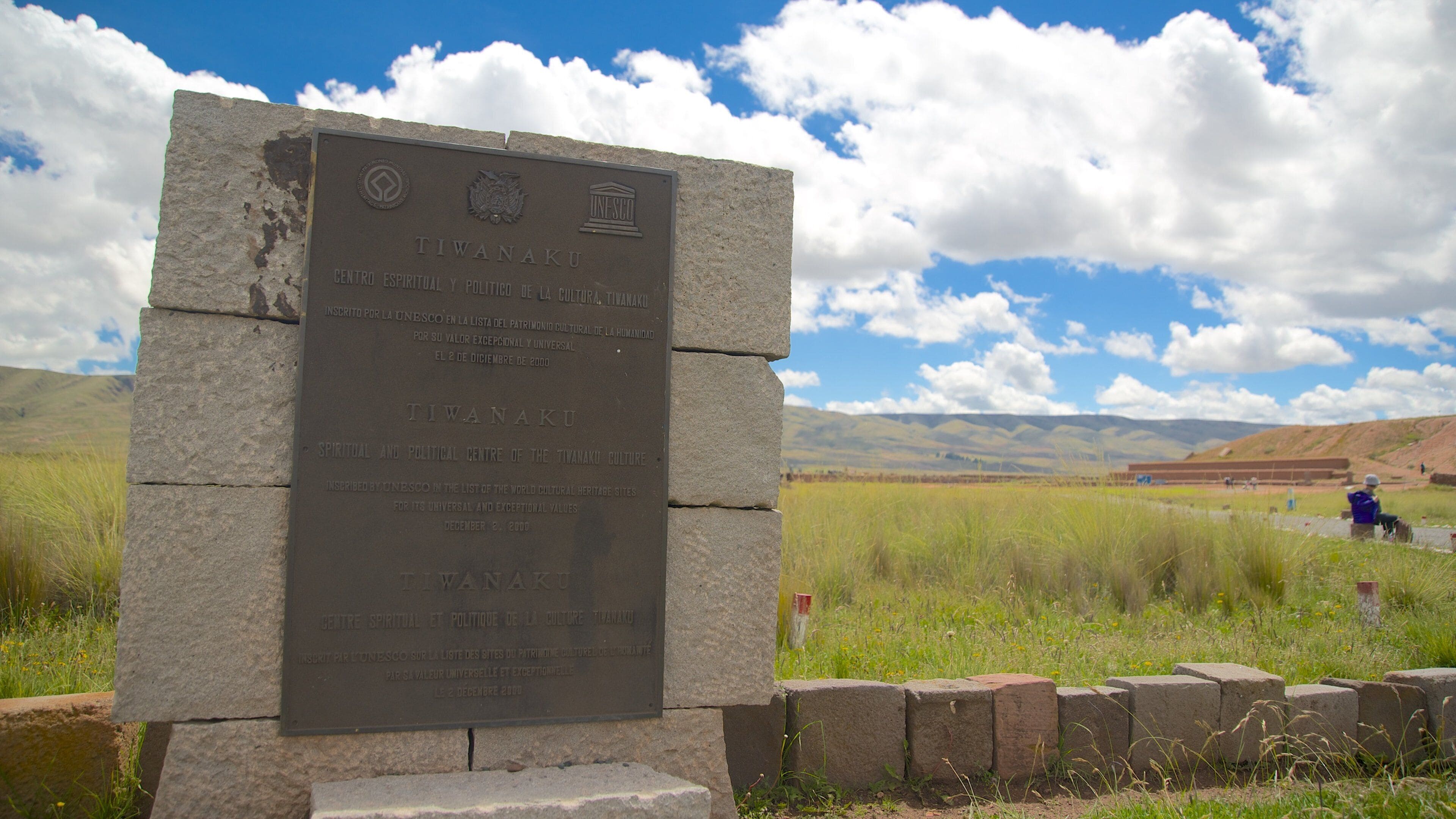 A stone plaque with inscriptions, set against a backdrop of a grassy field and distant mountains under a partly cloudy sky.