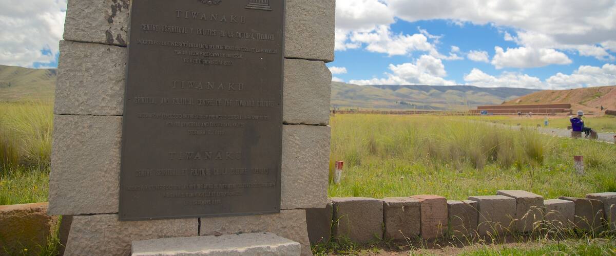 A stone plaque with inscriptions, set against a backdrop of a grassy field and distant mountains under a partly cloudy sky.