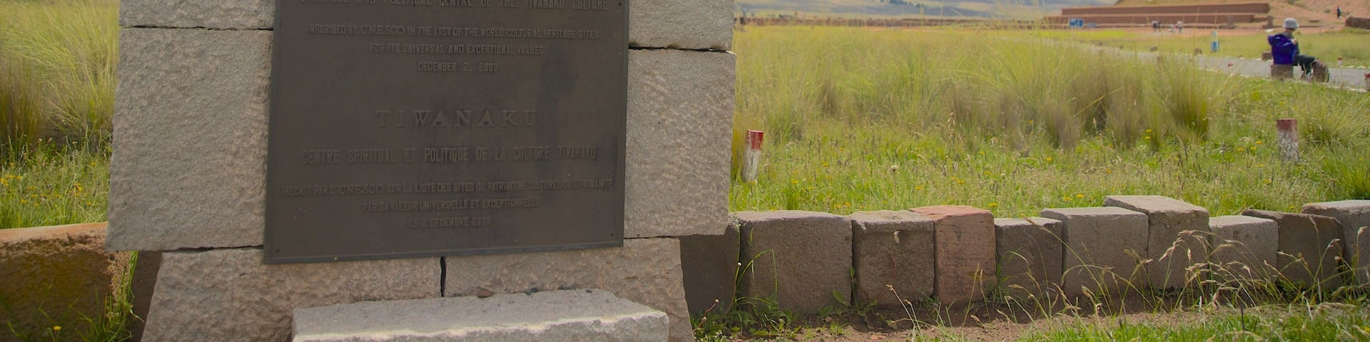 A stone plaque with inscriptions, set against a backdrop of a grassy field and distant mountains under a partly cloudy sky.