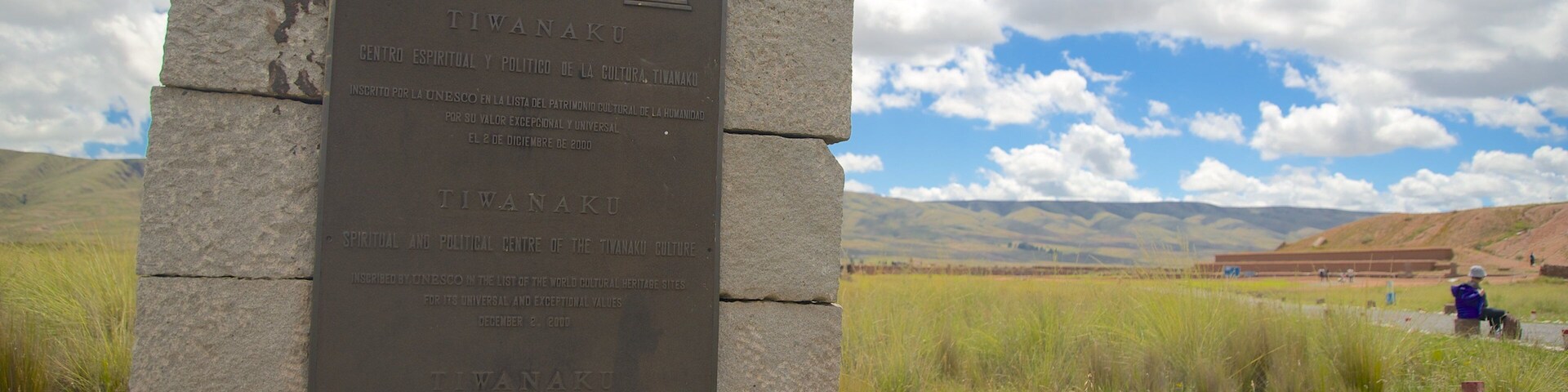Una placa de piedra con inscripciones, frente a un campo cubierto de hierba y montañas distantes bajo un cielo parcialmente nublado.