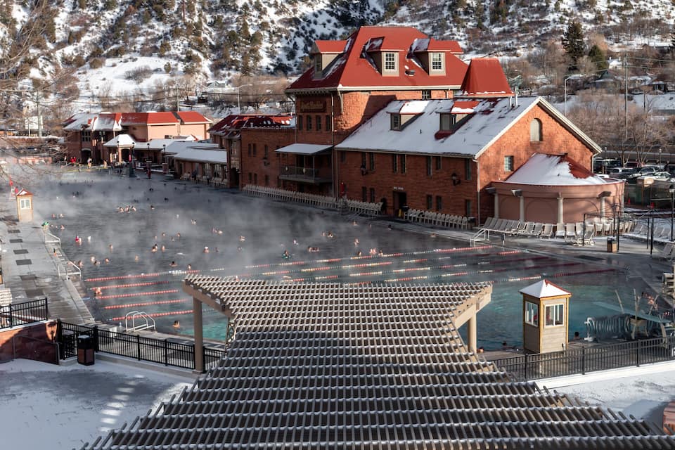 A snow-covered outdoor swimming area with a pool, a wooden gazebo, and a red-roofed building in the background.
