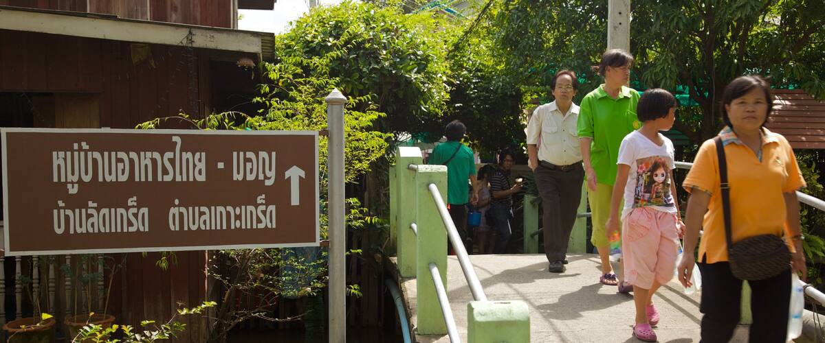 A sign with Thai text, a bridge, and people walking.