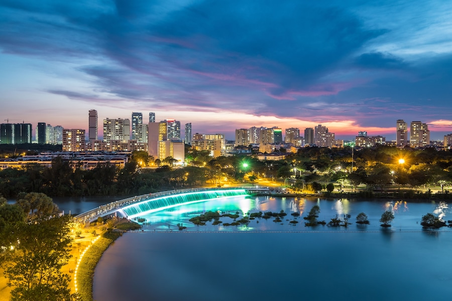 A city skyline at dusk with illuminated buildings, a waterfall, and a calm river reflecting the lights.