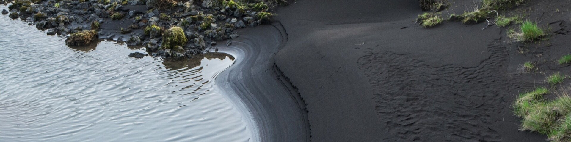 Orilla de río con arena oscura y vegetación verde.