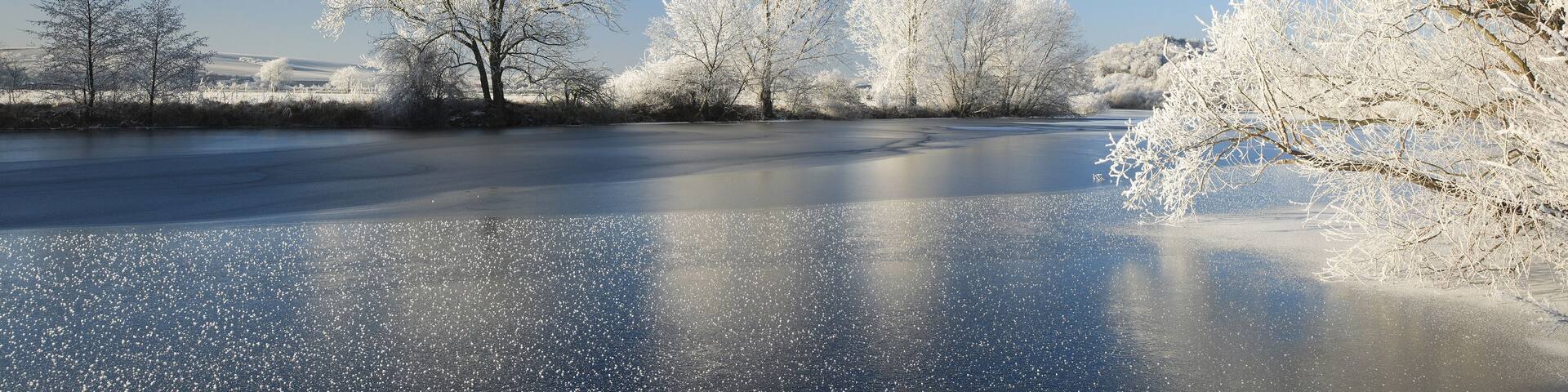 A frozen river with snow-covered trees reflecting on the ice.