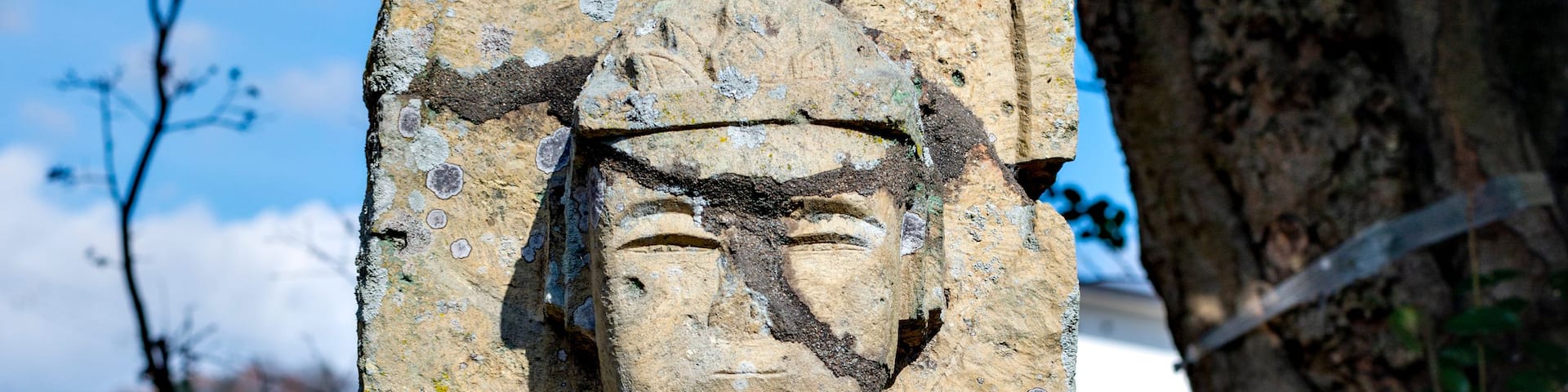 A stone statue with a circular motif at the top, set against a backdrop of trees and a clear sky.