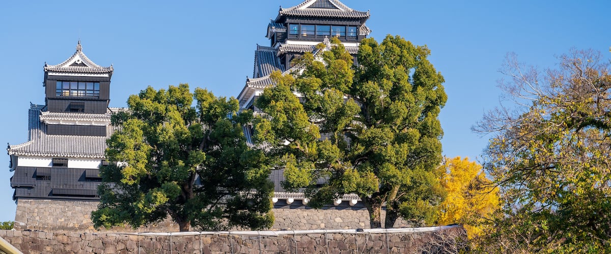 A traditional Japanese castle with multiple towers, surrounded by lush green trees and a clear blue sky.