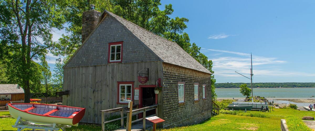 A wooden house with a red door and windows, a boat, and a lake in the background.