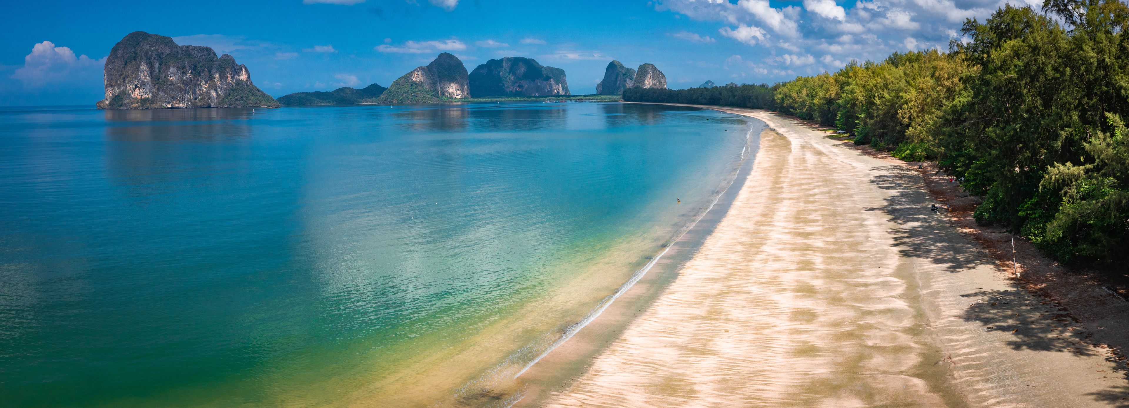 A coastal landscape with clear turquoise waters, a sandy beach, and a forested area with rocky cliffs in the background.