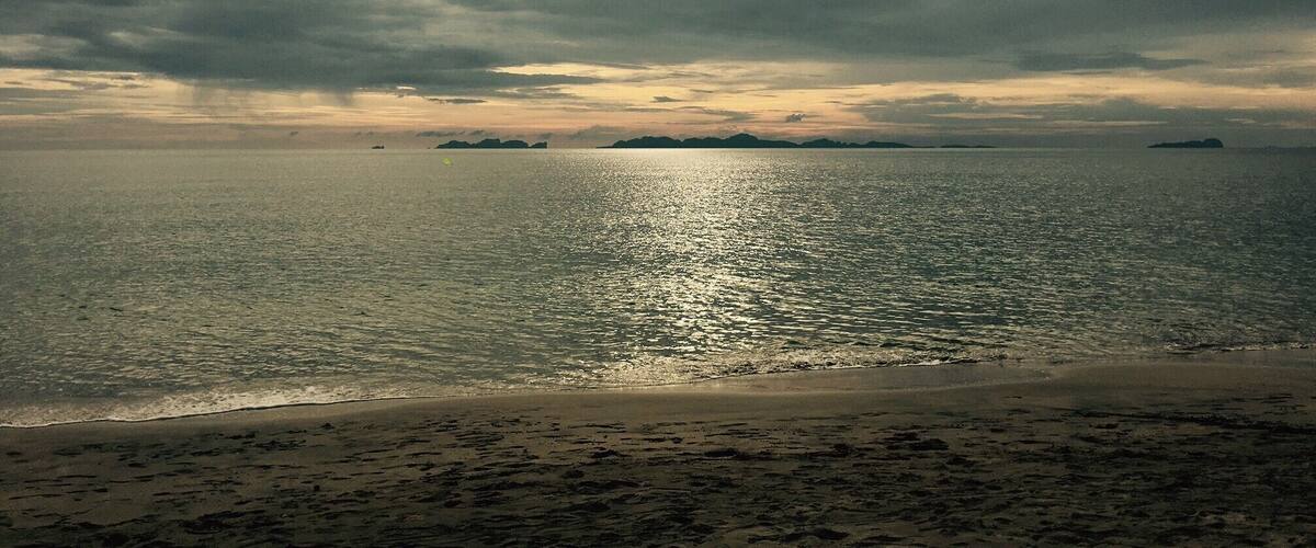 A beach with footprints leading to the water, a boat in the distance, and a cloudy sky.