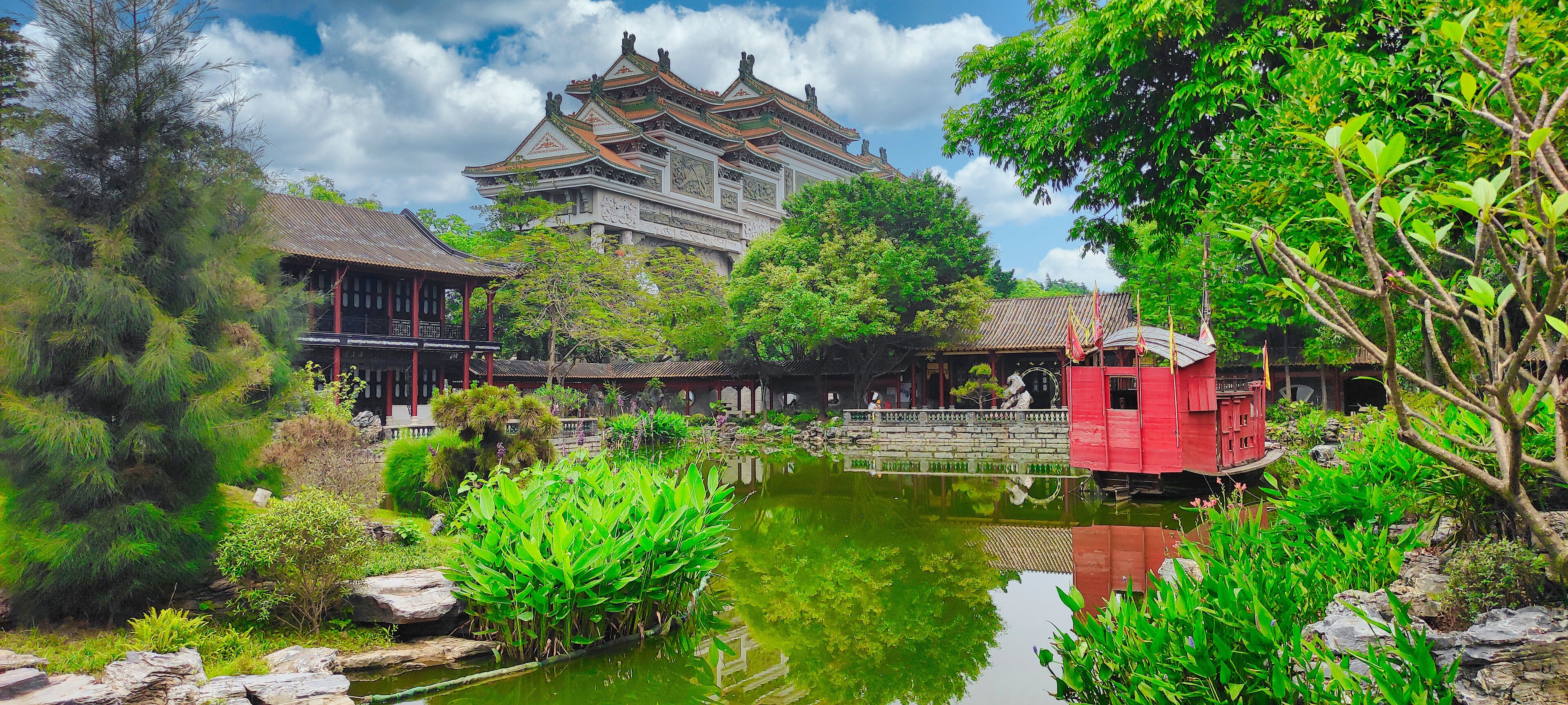 A traditional Chinese building with ornate architecture, a pond with water plants, and a red structure in the foreground.