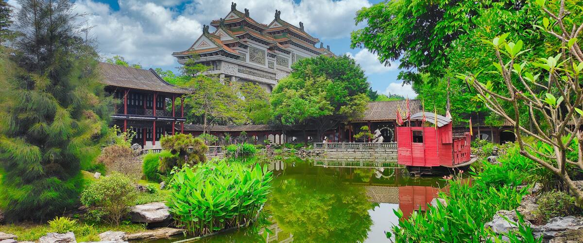 A traditional Chinese building with ornate architecture, a pond with water plants, and a red structure in the foreground.