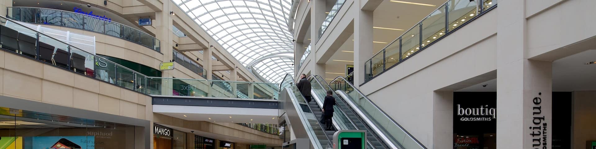 A multi-level shopping mall with escalators, glass railings, and various stores including a boutique.
