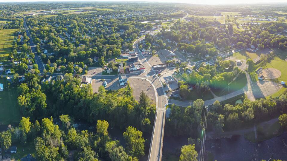 An aerial view of a small town with a central road, surrounding green areas, and a baseball field.