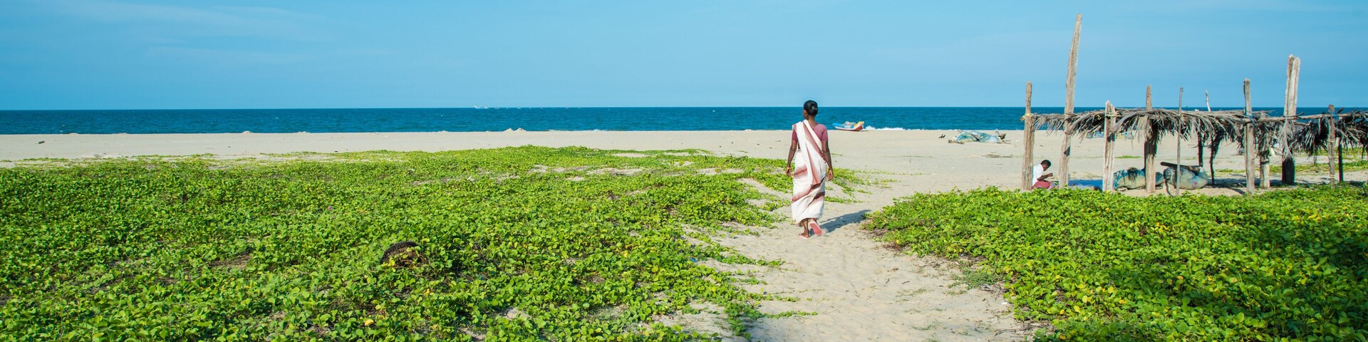 Una playa con una persona caminando por un sendero, vegetación verde y un cielo azul despejado.