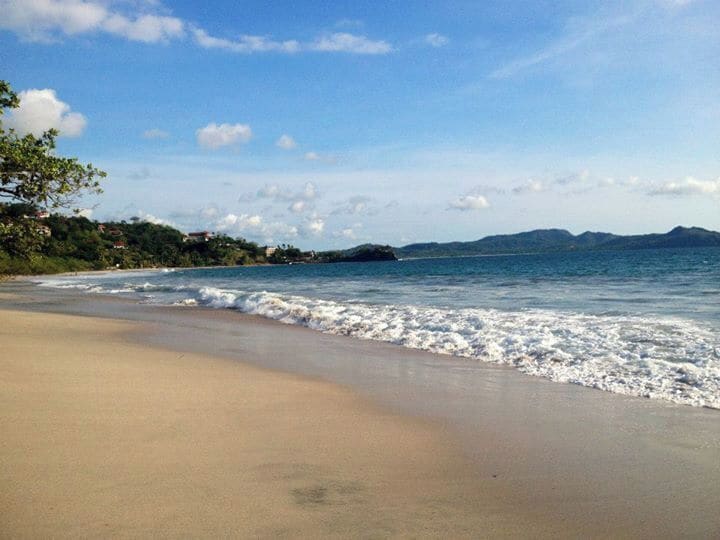 A sandy beach with gentle waves, a clear sky, and distant mountains.