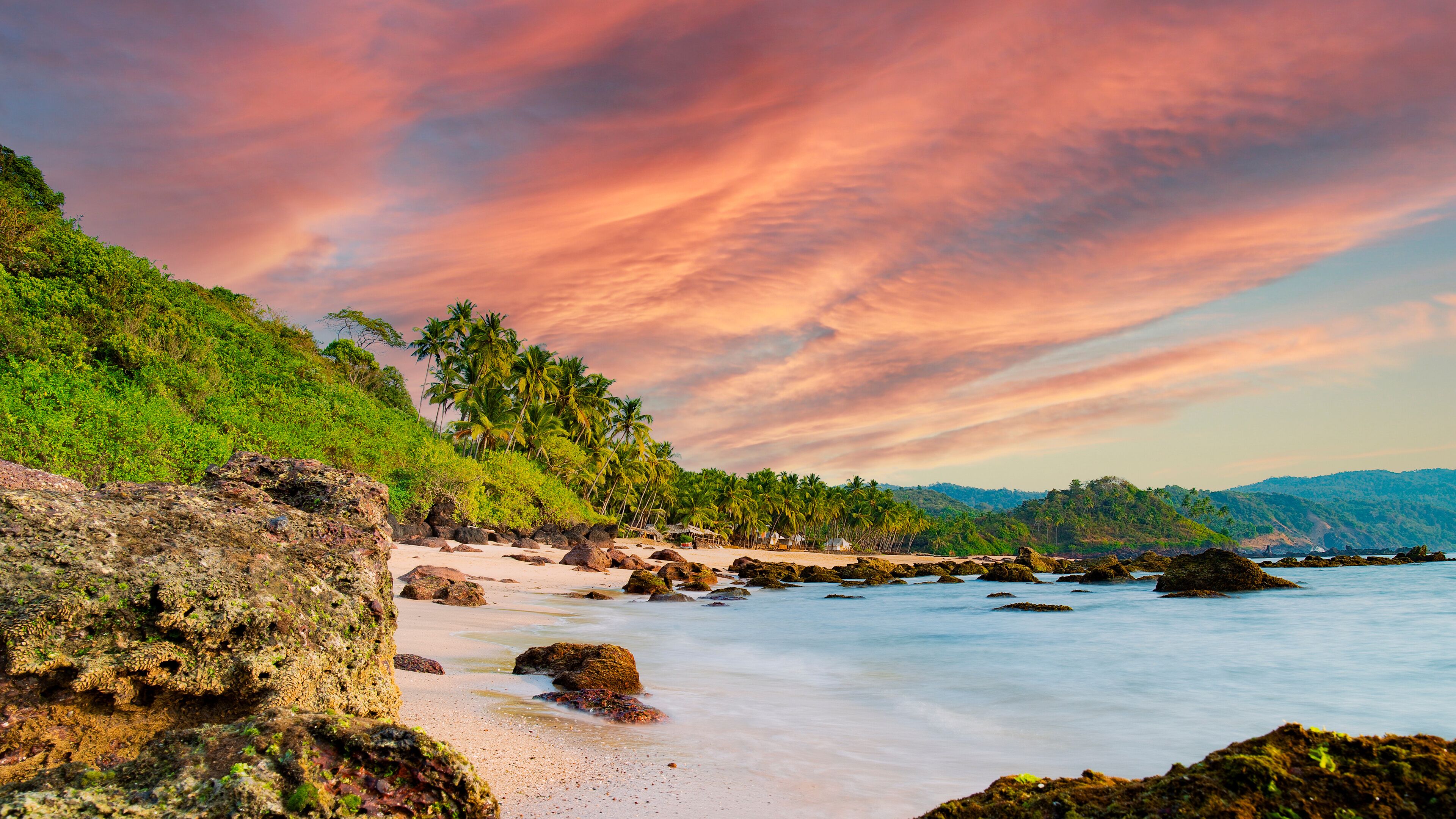 A tropical beach with palm trees, rocky shoreline, and a colorful sunset sky.