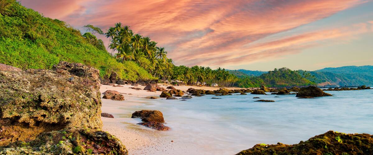 A tropical beach with palm trees, rocky shoreline, and a colorful sunset sky.