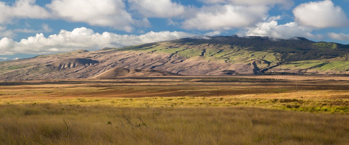 A vast open field with rolling hills and a mountain range in the background under a partly cloudy sky.