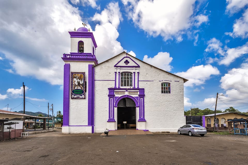 A church with a white facade and purple accents, a bell tower, and a clear blue sky with clouds.