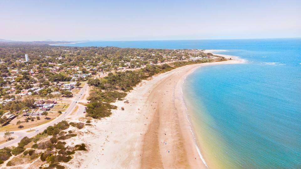 An aerial view of a coastal town with a long sandy beach, dense greenery, and a clear blue ocean.