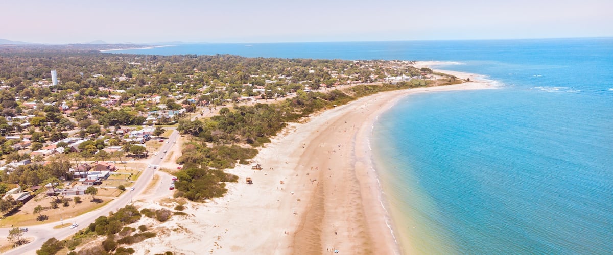 An aerial view of a coastal town with a long sandy beach, dense greenery, and a clear blue ocean.