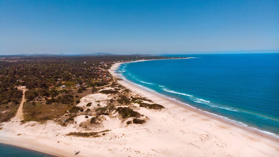 An aerial view of a coastal area with a sandy beach, a clear blue ocean, and a dense forested area with scattered buildings.
