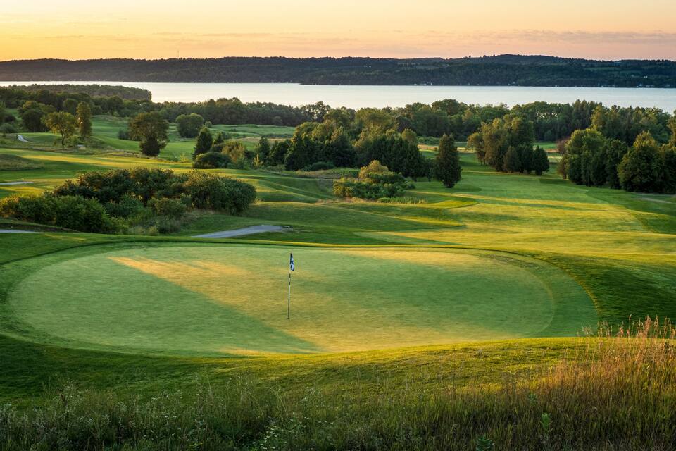 A golf course with a flagstick, surrounded by lush greenery and a lake in the background.