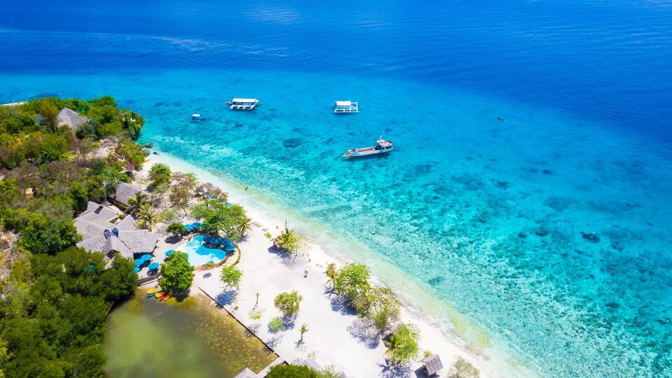 A tropical beach with clear turquoise waters, a small boat, and a resort with a pool.