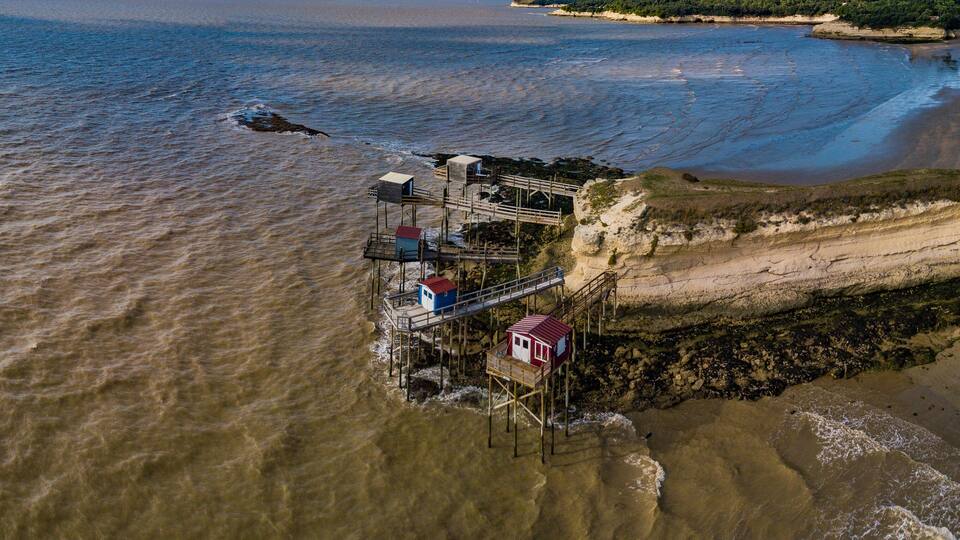 An aerial view of a coastal area with a wooden pier extending into the water, a beach, and a cliff with vegetation.