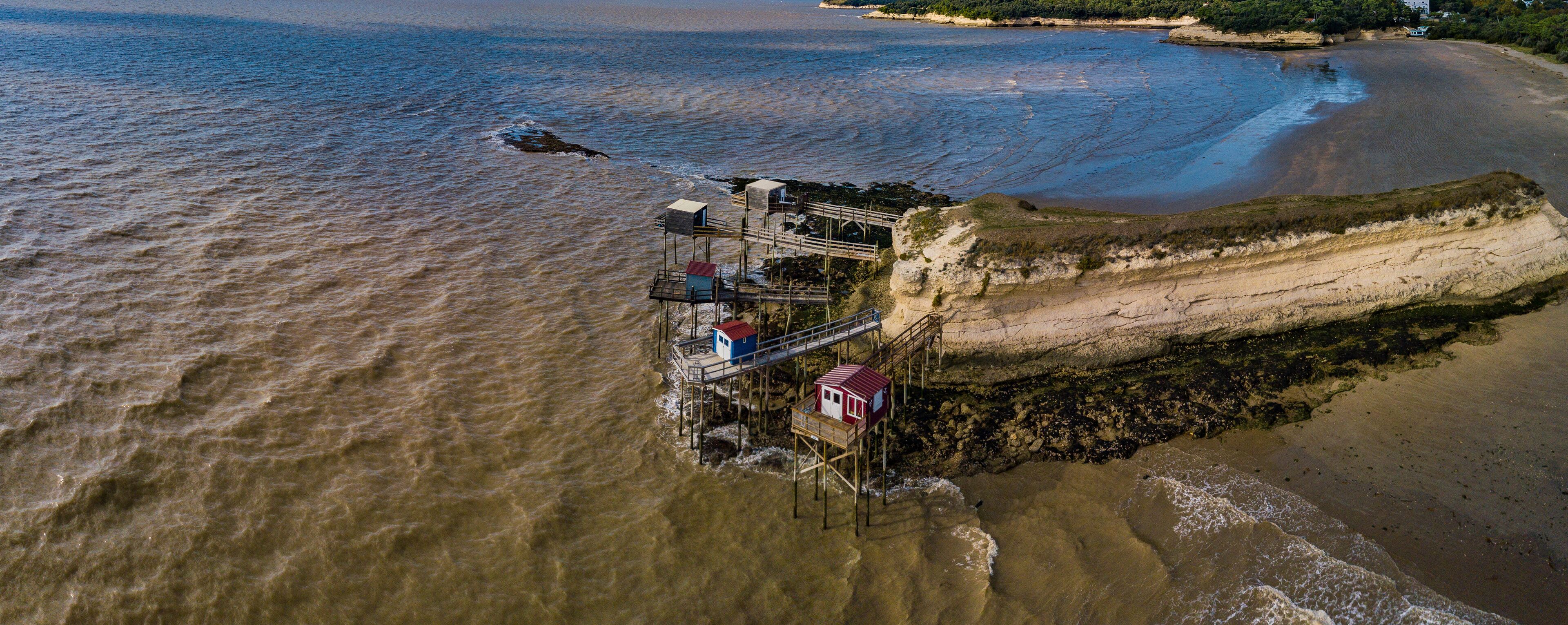 An aerial view of a coastal area with a wooden pier extending into the water, a beach, and a cliff with vegetation.