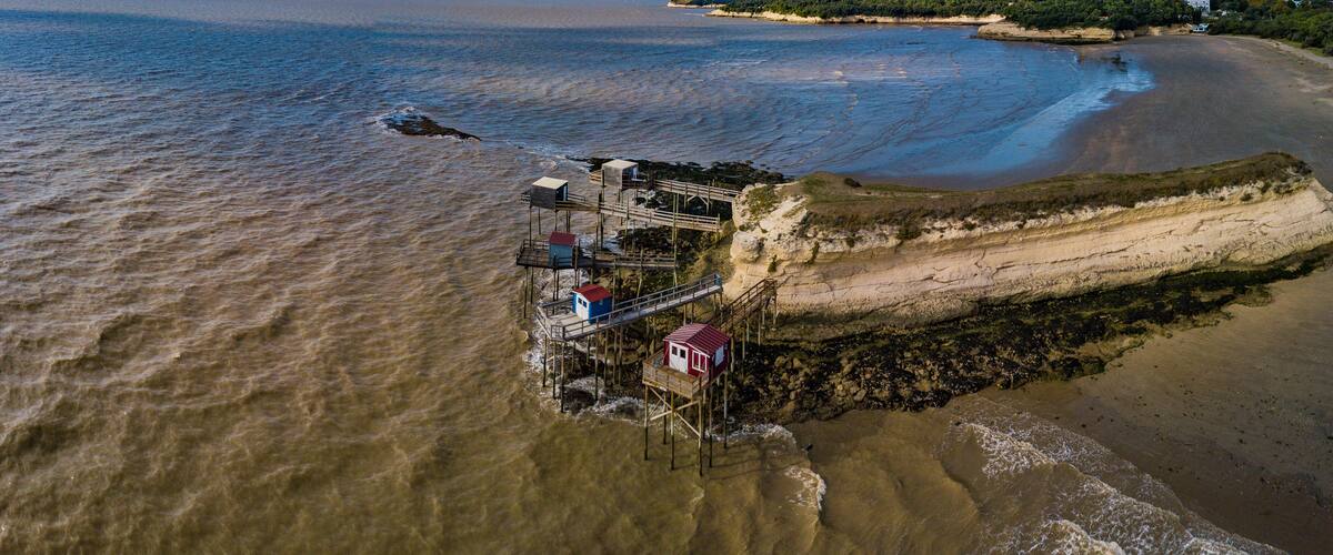 An aerial view of a coastal area with a wooden pier extending into the water, a beach, and a cliff with vegetation.