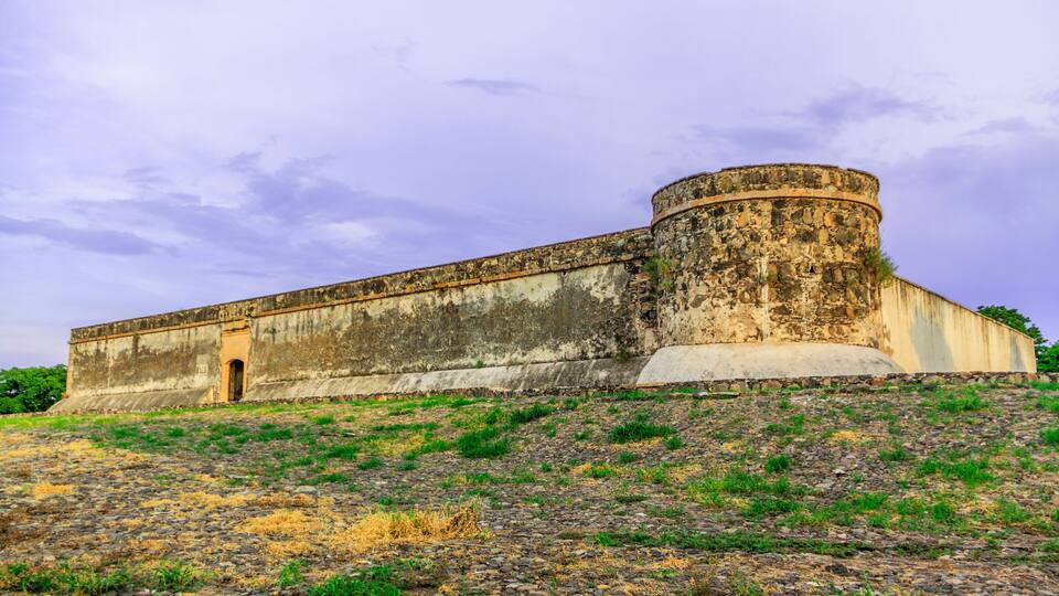 A large, circular fort with a flat roof, surrounded by a grassy hill with sparse vegetation.