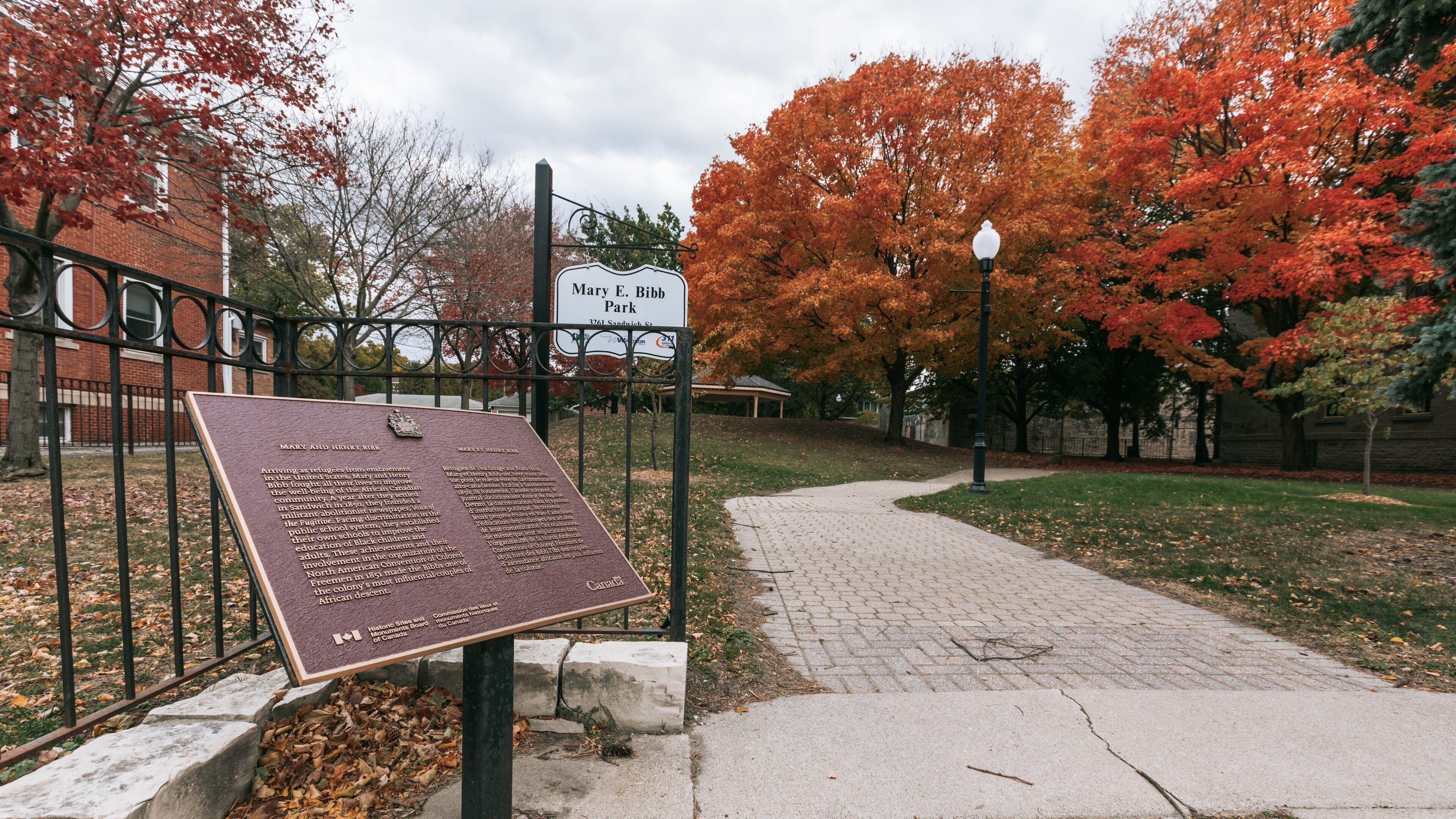 A park with a sign for Mary E. Bebb Park, surrounded by autumn trees and a brick building.