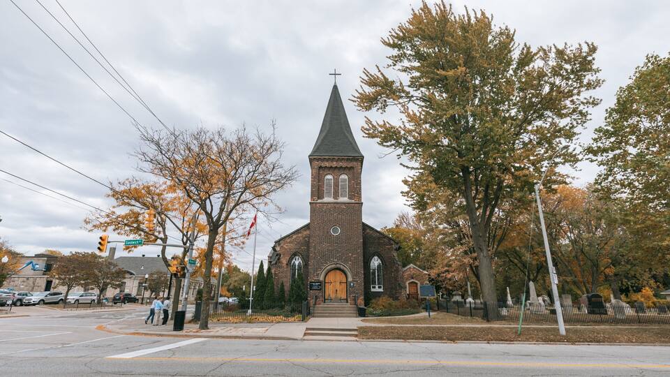 A brick church with a steeple and a cross on top, surrounded by autumn trees and a street with cars and pedestrians.