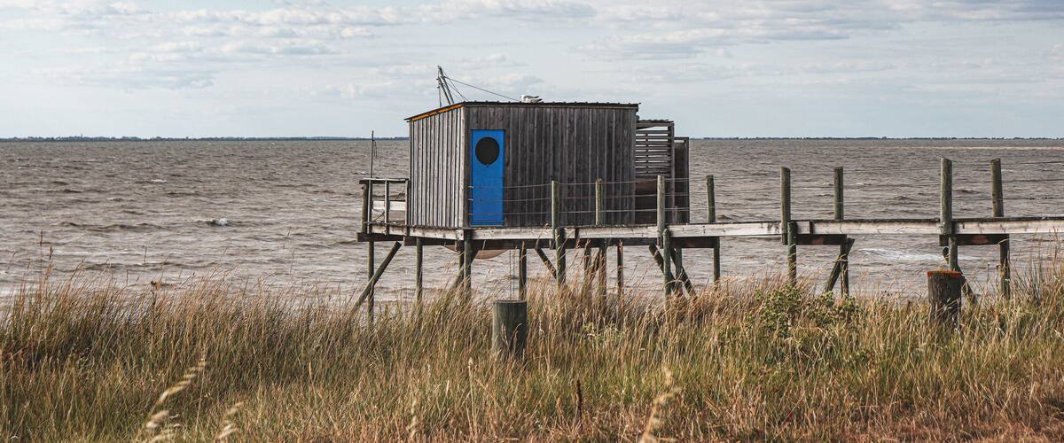 A wooden structure with a blue door on stilts over water, surrounded by grass and a cloudy sky.