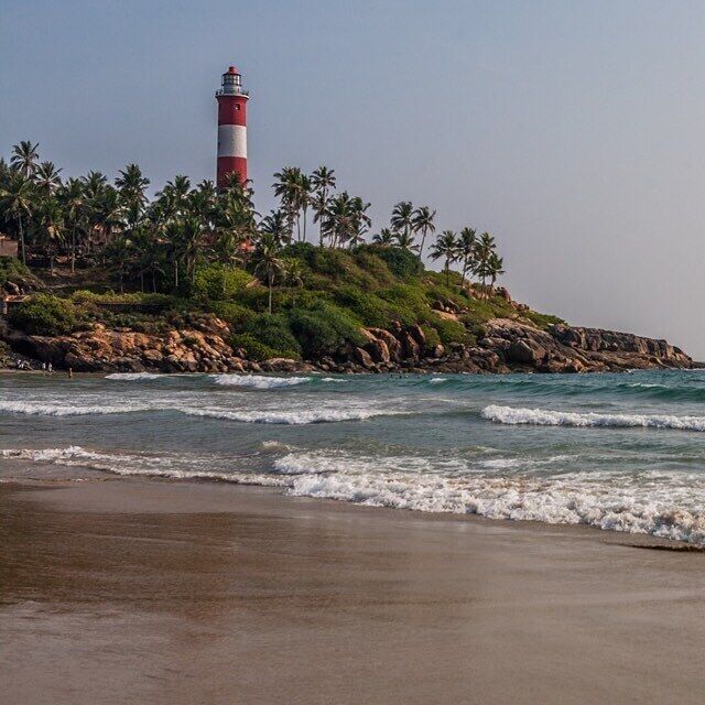 A lighthouse on a rocky outcrop with palm trees and waves crashing onto the shore.