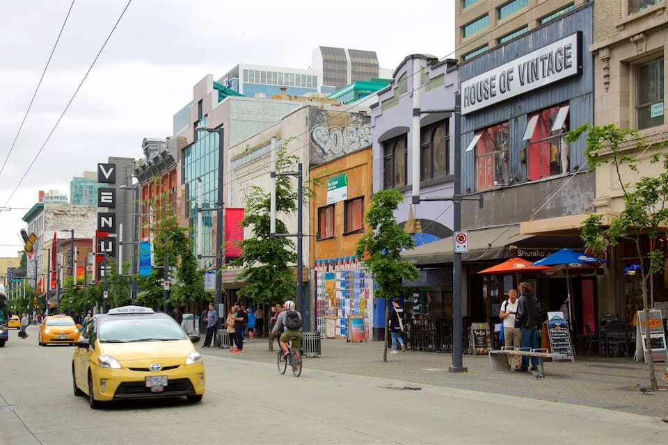 A busy urban street with a yellow taxi, cyclists, pedestrians, and various storefronts including a currency exchange.