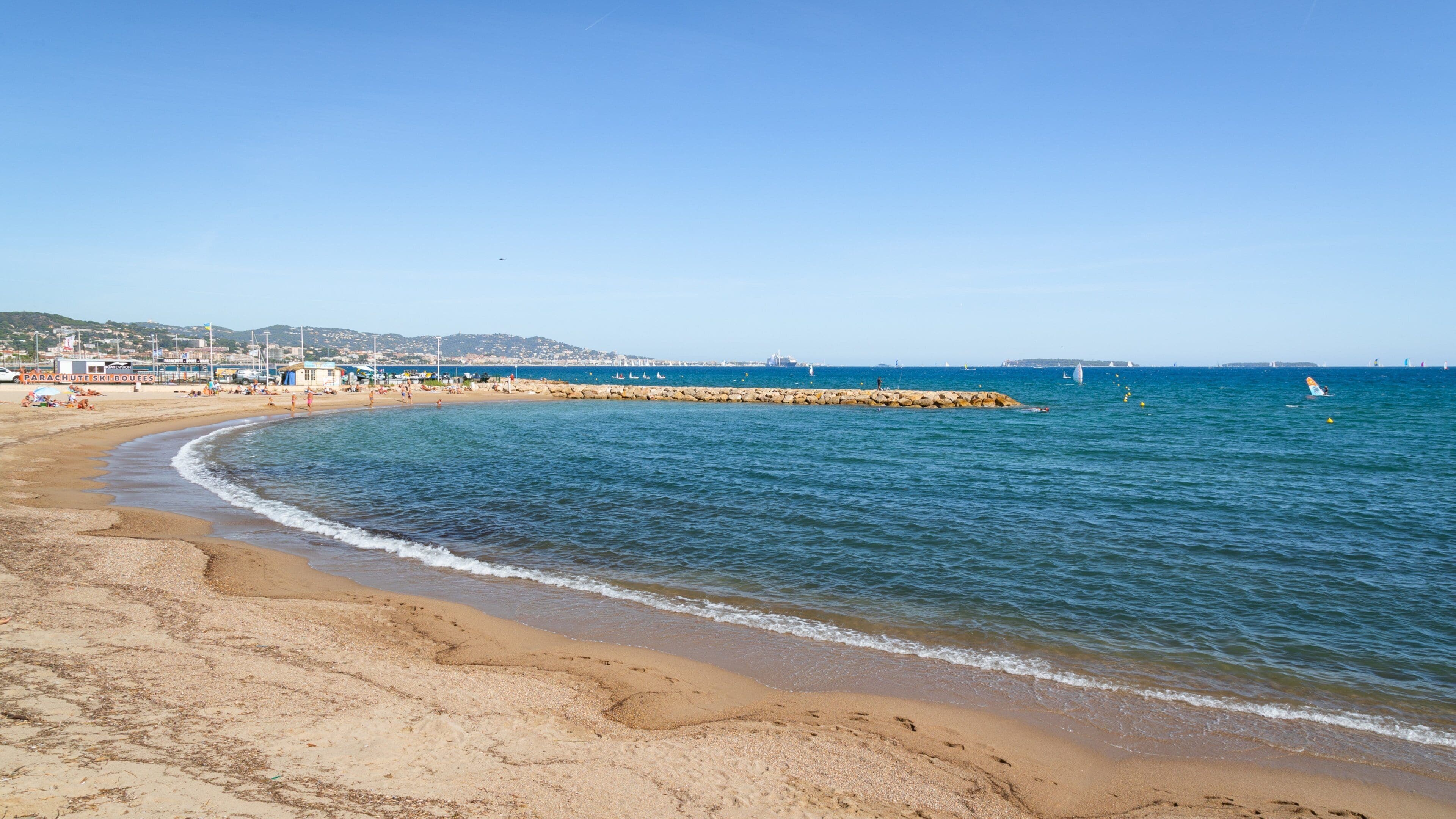 Une plage de sable fin bordée d’une eau d’un bleu limpide, une digue rocheuse et des bâtiments qui se dessinent à l’horizon.